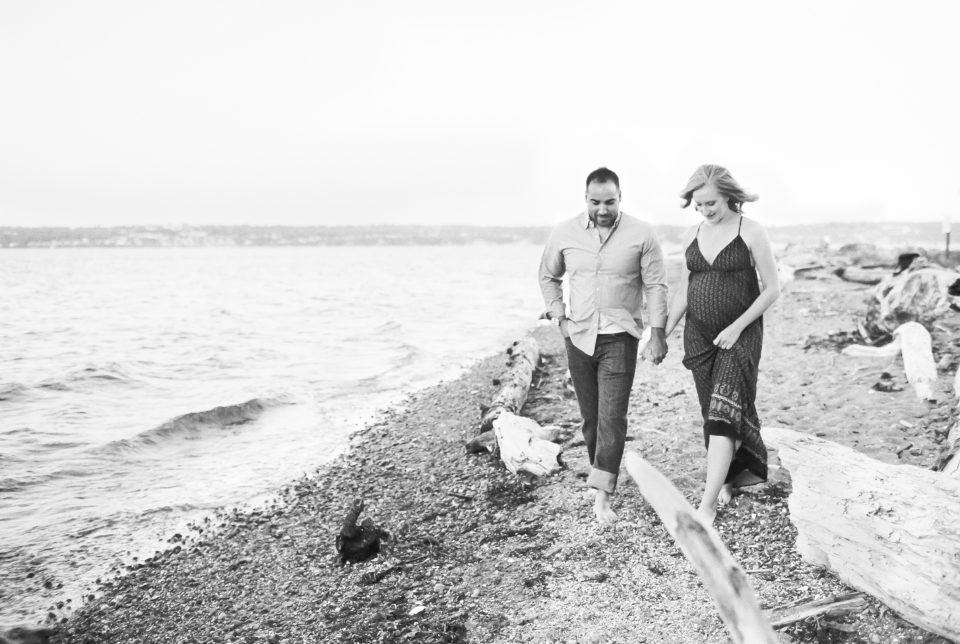 A pregnant woman holds her husbands hand as they walk along the beach