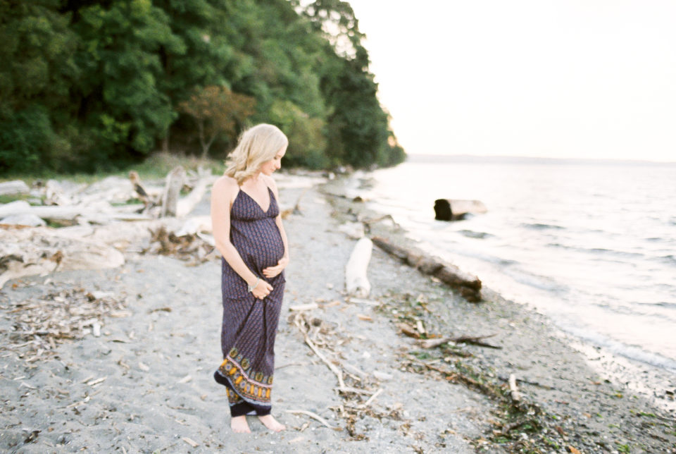 A pregnant woman in a blue dress holds her pregnant stomach on the beach