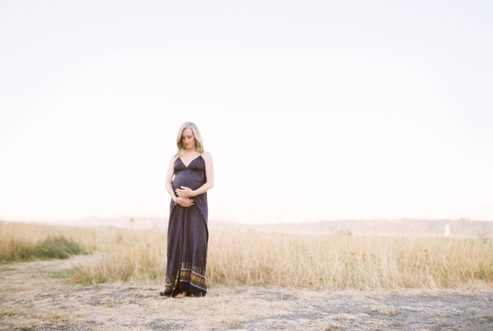 A blonde woman in a long dress holds her baby bump in a field