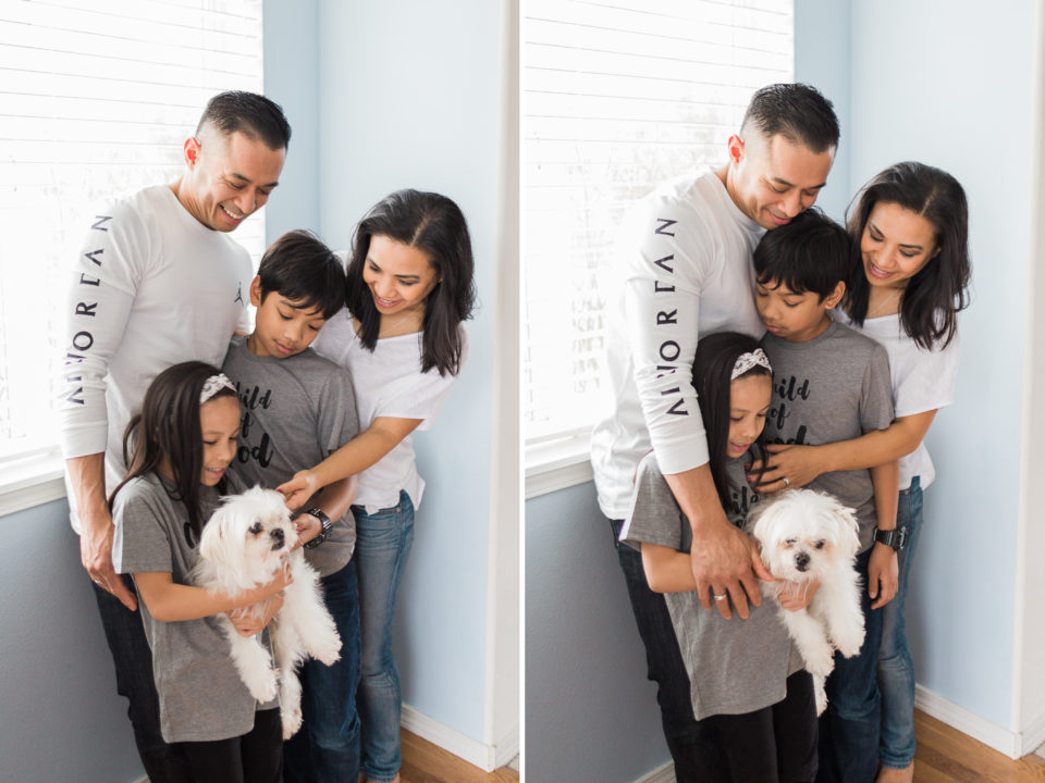 The family of four stand close together and pet their white pet dog 