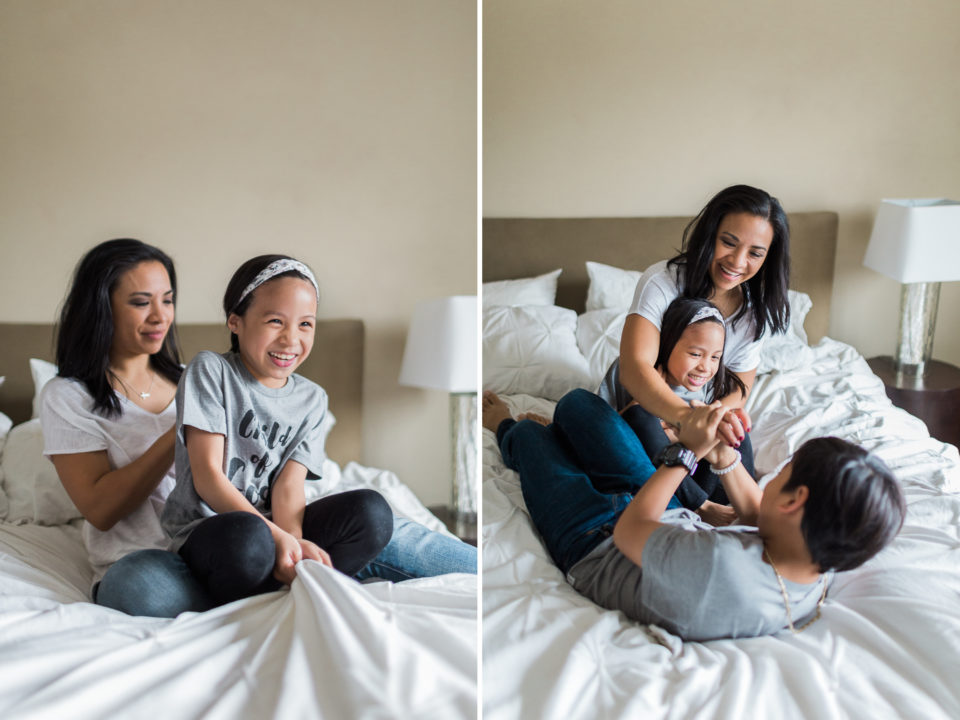 A mom braids her little girls hair as they sit on a white bed together