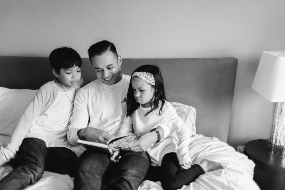 A dad wraps his arms around his young kids and reads them a book