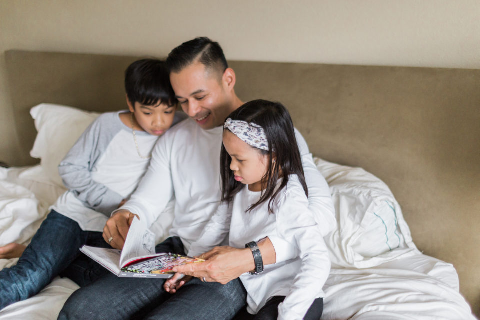 Two kids in white shirts listen to their dad read them a book