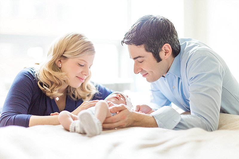 The parents of a newborn baby boy smile at him in front of a bright and airy window