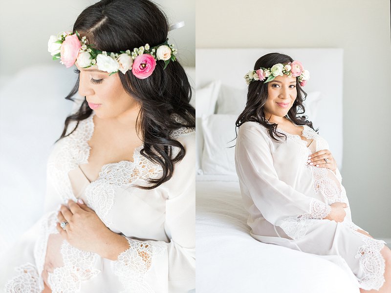 A pregnant mother sits on a white bed cradling her baby bump with a pink flower crown in her hair