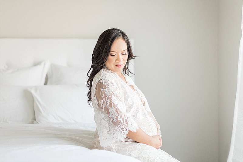A gorgeous expectant mama with dark hair sits on a white bed and smiles at her bump