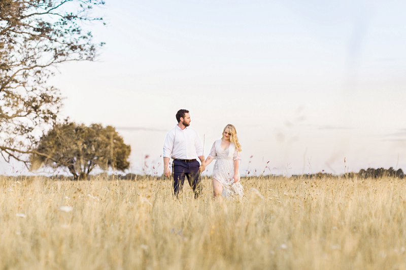 The couple stands in a field of tall golden grass and holds hands