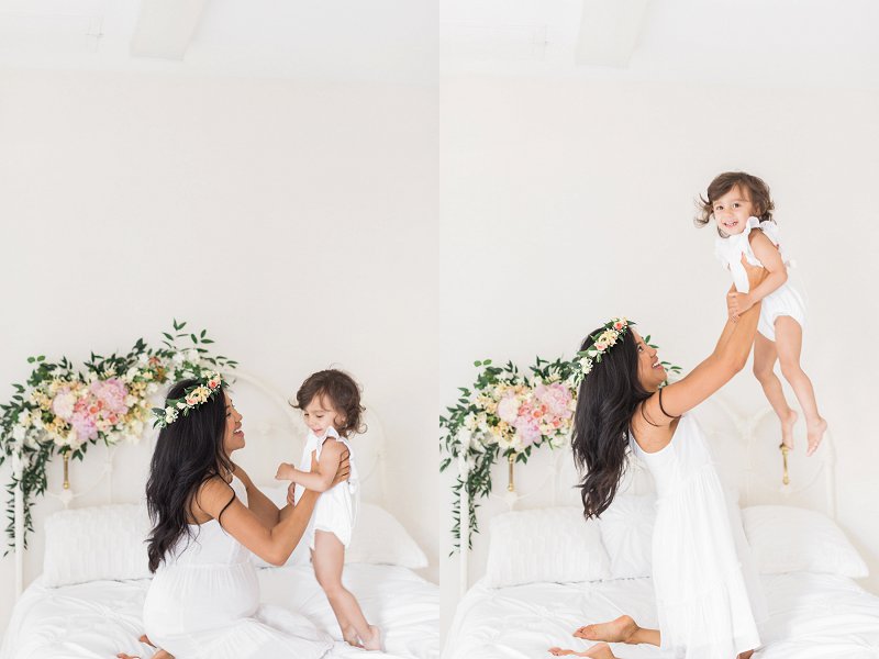 The cute toddler smiles with glee as her mother bounces her on the bed