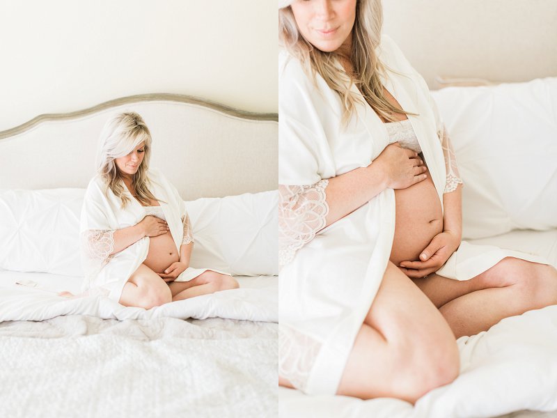 A blonde woman wearing a cream robe sits on her bed and cradles her baby bump with a small smile