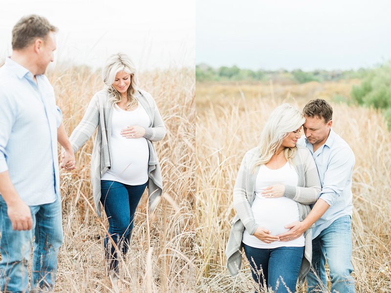 Standing in a field of tall golden grass a couple stands close together and smiles down at her growing baby