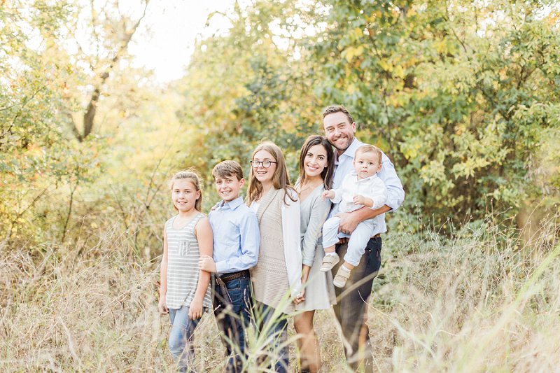 A family standing from tallest to shortest holds each other close and smiles at their photographer with tall grass surrounding them