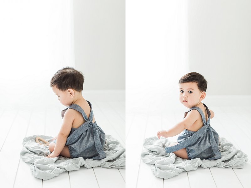 A sweet baby sitting on a blanket on the white floor looks over his shoulder at the camera