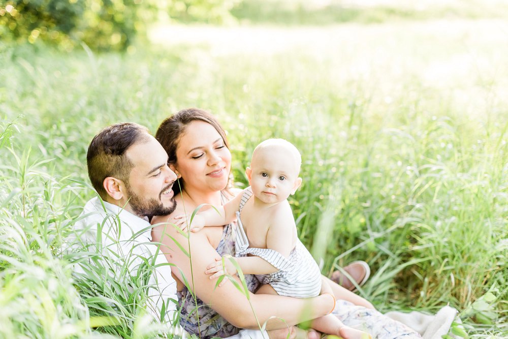 The family cuddle together in a field of tall grass while the the little boy looks at the camera
