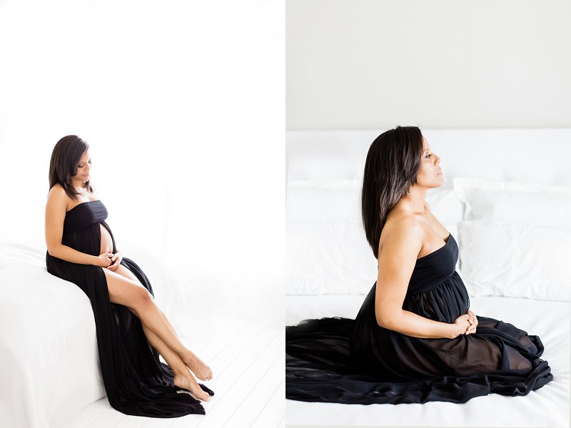 The expectant mother wearing a long black dress sits at the edge of a bed and holds her pregnant bump