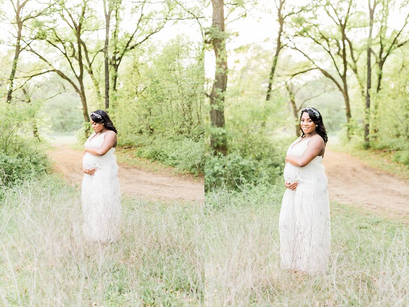 As she stands in a small area of tall grass in the forest, a pregnant woman smiles down at her baby bump