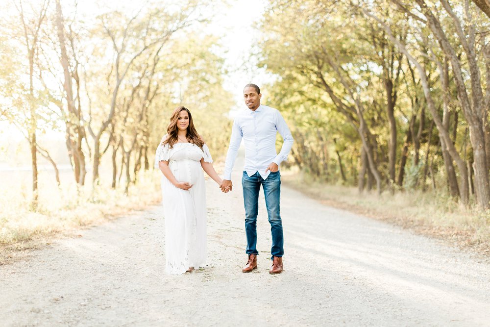 The soon to be parents stand side by side on a gravel path way and smile at the camera 