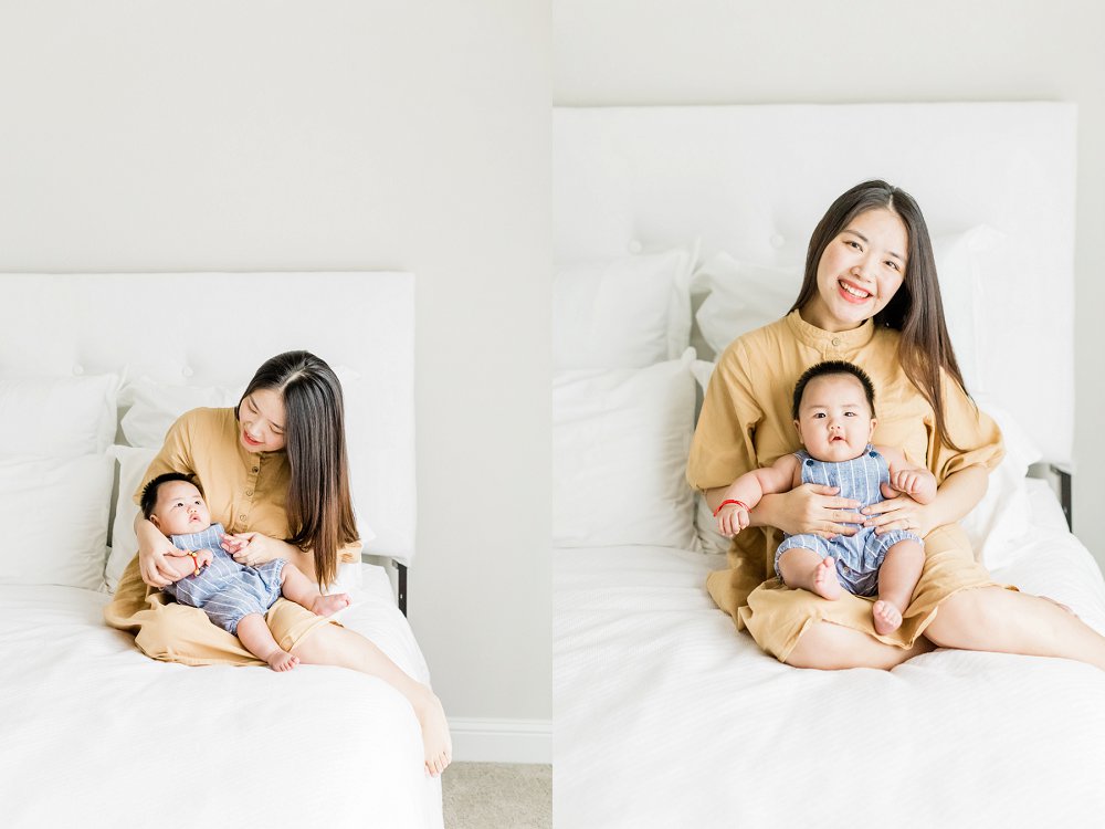 Mom and Baby sit together on a cozy white bed and smile and laugh together