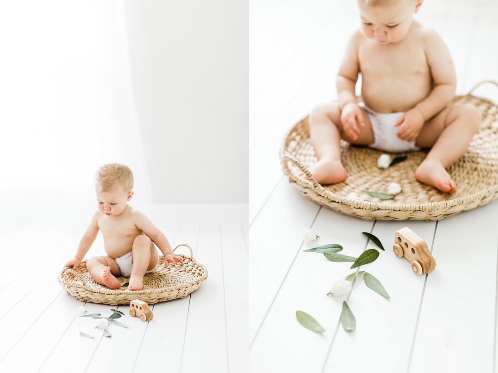 A baby sitting in a rattan basket looks around at the leaves scattered on the floor around him