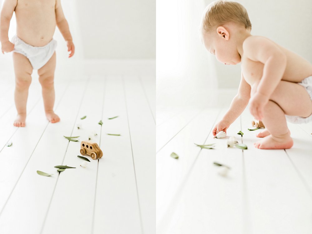 A chubby baby bends down to pick up a leaf from the bunch scattered on the white studio floor
