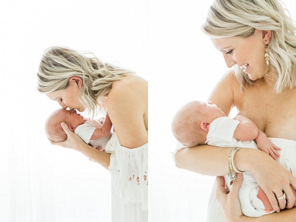 Mom wearing white dress kissing and smiling at newborn baby wearing a white onesie
