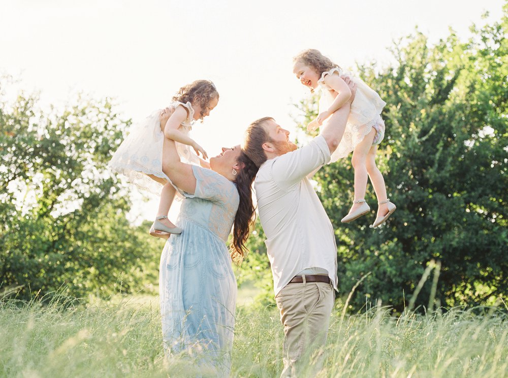Mom and Dad hold their girls up high among the trees with the sun shining behind them
