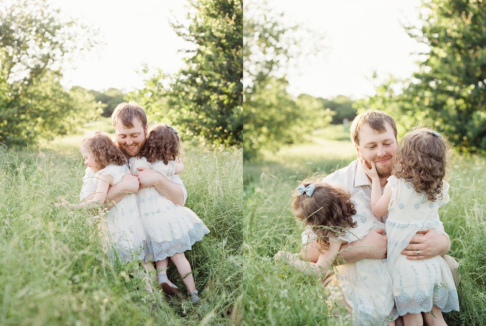 Two twin girls with brown ringlet curls give their Dad a big hug while he crouches on the ground